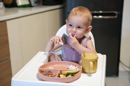 Portrait of little toddler girl sitting in a high chair in the kitchen eating lunch. She thoughtfully puts rice in her mouth with her hand and learns to eat with utensils. The baby eats independently.の写真素材