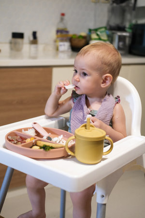 A little girl toddler is sitting on a high chair and eating a cracker. She plays with the cracker imagining that it is an airplane that flies into her mouth and bites is off.の写真素材