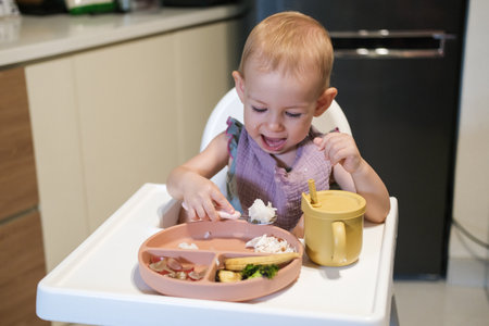 A little toddler girl sits in a high chair and eats her lunch. The baby is learning to eat with a fork, but she picks up rice with her hand and puts it in her mouth. She plays with food and has fun.の写真素材