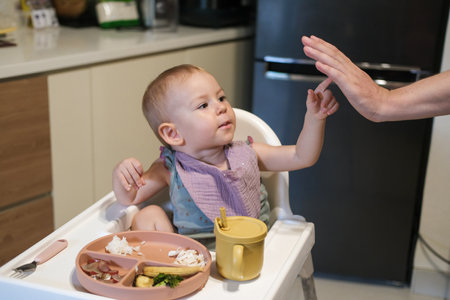 A little toddler girl sits in a high chair and eats her lunch. The baby is learning to eat with a fork, but she picks up rice with her hand and puts it in her mouth. She plays with food and has fun.の写真素材