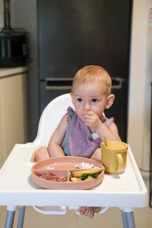 Portrait of little toddler girl sitting in a high chair in the kitchen eating lunch. She thoughtfully puts rice in her mouth with her hand and learns to eat with utensils. The baby eats independently.の写真素材