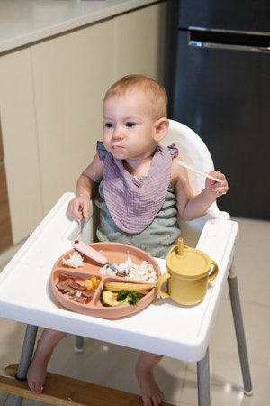 A little girl toddler is sitting on a high chair and eating a cracker. She plays with the cracker imagining that it is an airplane that flies into her mouth and bites is off.の写真素材