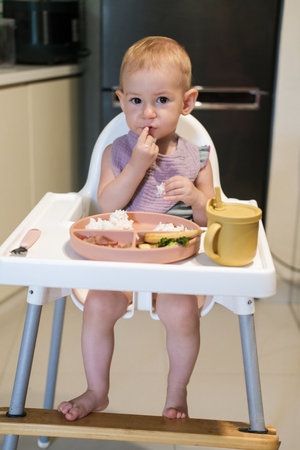 Portrait of little toddler girl sitting in a high chair in the kitchen eating lunch. She thoughtfully puts rice in her mouth with her hand and learns to eat with utensils. The baby eats independently.の写真素材