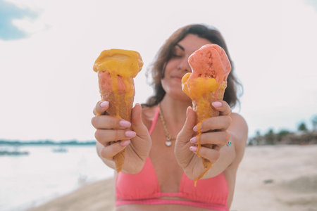 Portrait of a young woman holding two ice cream cones on the beach in a swimsuit. Ice cream runs down her hand and quickly melts. Refreshing summer dessert.の写真素材