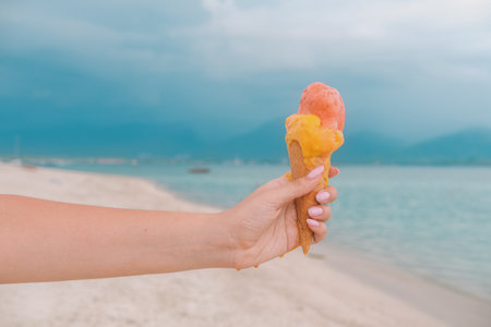 Close up of woman hand holding waffle cone with ice cream melting and running down her hand against blue ocean on beach. Ice cream running and dripping on your hand. Extreme heat. Summer background.の写真素材