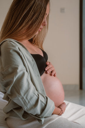 Side shot of pregnant woman belly She is sitting on bed in her bedroom and gently stroking her round belly in anticipation of the baby arrival. Third trimester of pregnancy, health care and childbirthの写真素材