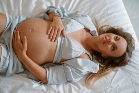 Portrait of a pregnant young woman. She lies on the bed in her pajamas and strokes her round belly in anticipation of the arrival of the baby. Third trimester of pregnancy, medical care and childbirthの写真素材