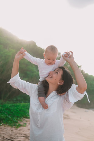 Portrait of mother and baby who sits on her neck and looks down. The woman smiles and walks with baby on the beach near the ocean. Vacation with little son. Mothers Day.の写真素材