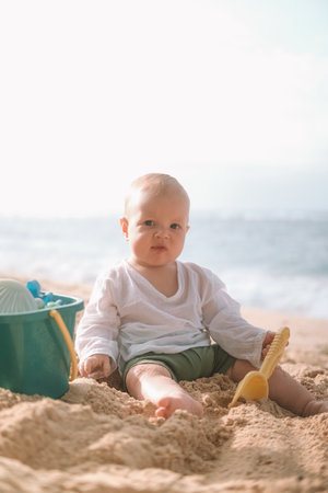 Portrait of a baby sitting on the beach on sand and playing with toys, a shovel and a bucket. Children entertainment on vacation on the beach.の写真素材