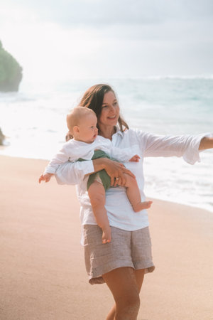Portrait of a young woman walking on the beach with a baby in her arms. She hugs and kisses him on the beach with the ocean in the background. Child first vacation with parents. Mothers Day.の写真素材