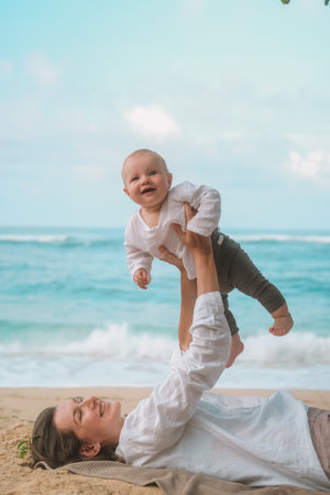 Portrait of woman lying on the sand with ocean in background and holding her baby up in her arms and playing with him and kissing him. Mother and child laughing and having fun on vacation. Mothers Dayの写真素材
