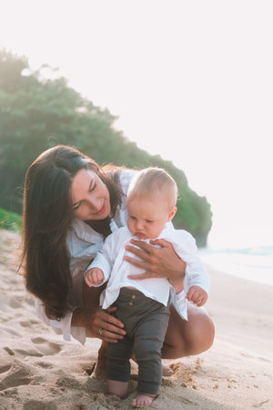Portrait of mother and baby with blue eyes taking first steps on the beach by the ocean. He looks forward with curiosity and laughs. Mother holds her sons hands and helps him. Mothers Day.の写真素材