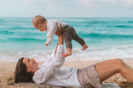 Portrait of woman lying on the sand with ocean in background and holding her baby up in her arms and playing with him and kissing him. Mother and child laughing and having fun on vacation. Mothers Dayの写真素材