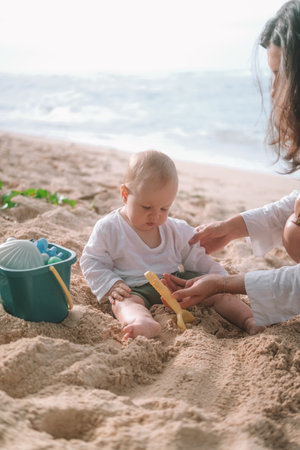 Portrait of a baby sitting on the beach on sand and playing with toys, a shovel and a bucket. Children entertainment on vacation on the beach. Next to him sits his mother and helps him. Mothers Day.の写真素材