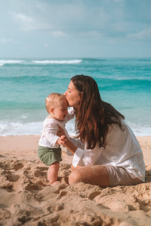 Portrait of mother and baby on vacation on the beach by the ocean. The little boy is capricious and crying, mother calms him down kisses and tickles him to cheer him up. Mothers Day. Travel with childの写真素材