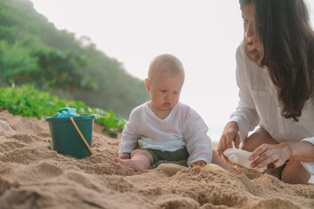 Portrait of a baby sitting on the beach on sand and playing with toys, a shovel and a bucket. Children entertainment on vacation on the beach. Next to him sits his mother and helps him. Mothers Day.の写真素材