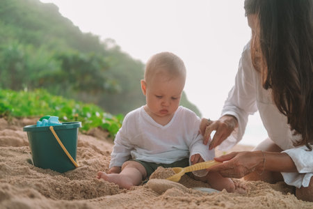 Portrait of a baby sitting on the beach on sand and playing with toys, a shovel and a bucket. Children entertainment on vacation on the beach. Next to him sits his mother and helps him. Mothers Day.の写真素材