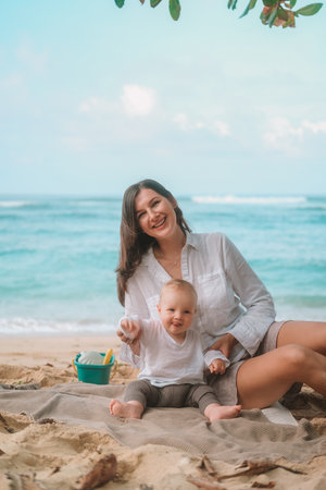 Portrait of a baby sitting on the beach on sand and playing with toys, a shovel and a bucket. Children entertainment on vacation on the beach. Next to him sits his mother and helps him. Mothers Day.の写真素材