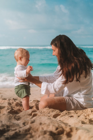 Portrait of mother and baby on vacation on the beach by the ocean. The little boy is capricious and crying, mother calms him down kisses and tickles him to cheer him up. Mothers Day. Travel with childの写真素材