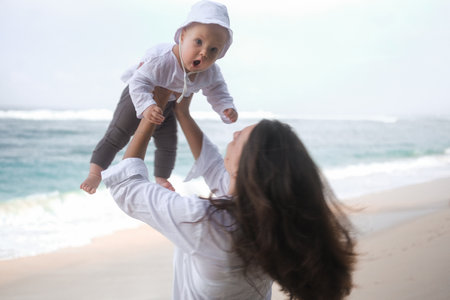 Portrait of a young woman walking on the beach with a baby in her arms. She throws the baby up in the air with the ocean in the background, the baby is surprised. Child first vacation with parents.の写真素材