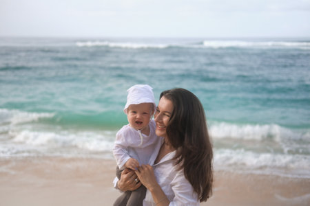 Portrait of a young woman holding a baby. She hugs and kisses him on the beach with the ocean in the background. Childs first vacation with parents. Mothers Day. Love for her son.の写真素材