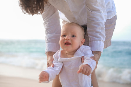Portrait of a baby with blue eyes taking his first steps on the beach by the ocean. He looks forward curiously and laughs. Mom holds her sons hands and helps him. Mothers Day. Moms love.の写真素材