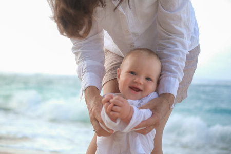 Portrait of a baby with blue eyes taking his first steps on the beach by the ocean. He looks forward curiously and laughs. Mom holds her sons hands and helps him. Mothers Day. Moms love.の写真素材