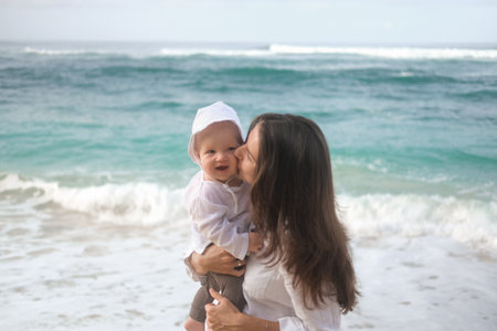 Portrait of a young woman holding a baby. She hugs and kisses him on the beach with the ocean in the background. Childs first vacation with parents. Mothers Day. Love for her son.の写真素材