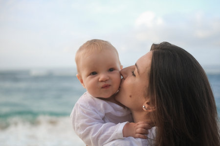 Close-up portrait of a baby with blue eyes being held by his mother against ocean. The mother is kissing her son on the cheek, the child is laughing. Vacation with a child. Mothers Day. Family travel.の写真素材
