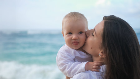 Close-up portrait of a baby with blue eyes being held by his mother against ocean. The mother is kissing her son on the cheek, the child is laughing. Vacation with a child. Mothers Day. Family travel.の写真素材