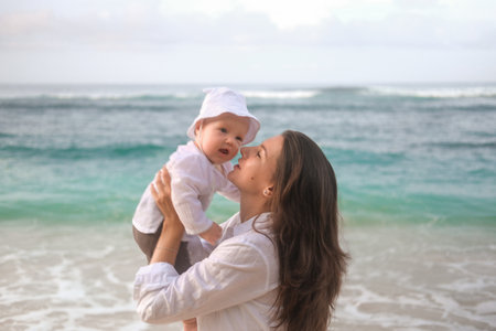 Portrait of a young woman holding a baby. She hugs and kisses him on the beach with the ocean in the background. Childs first vacation with parents. Mothers Day. Love for her son.の写真素材