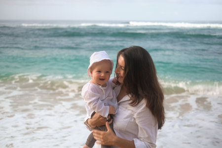 Portrait of a young woman holding a baby. She hugs and kisses him on the beach with the ocean in the background. Childs first vacation with parents. Mothers Day. Love for her son.の写真素材