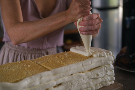 Close up of hands of young woman pastry chef preparing big birthday cake in home kitchen. She mixes...