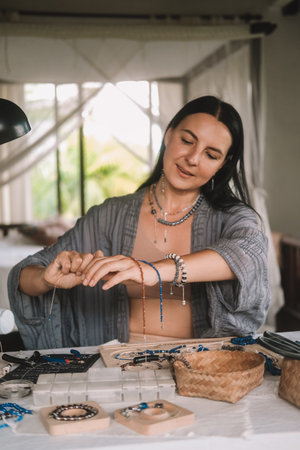 Portrait of a young brunette woman jewelry designer. She is sitting at a work table on which lie tools and various beads from which she creates jewelry. Home small business, startup. Creative hobby.の写真素材