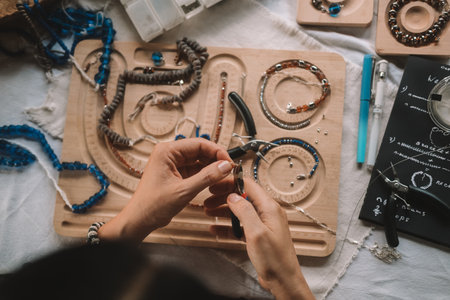 Close up of hands woman jewelry designer. She is sitting at a workspace with tools on it and is...