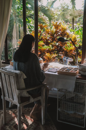 Back view of a young brunette woman, a jewelry designer. She is sitting at her workspace, where she has tools and various beads that she uses to create jewelry. Home based small business, startup.の写真素材