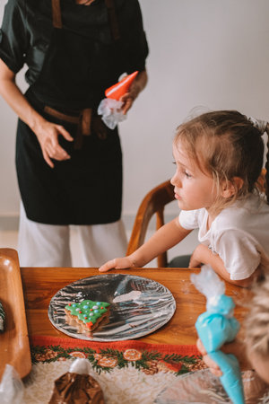 A female pastry chef teaches children how to decorate Christmas gingerbread. She holds a pastry bag and draws on the gingerbread, demonstrating to the children. Festive workshop for childrenの写真素材