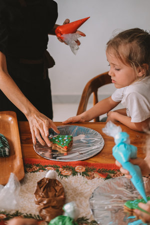 A female pastry chef teaches children how to decorate Christmas gingerbread. She holds a pastry bag and draws on the gingerbread, demonstrating to the children. Festive workshop for childrenの写真素材
