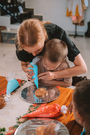 A female pastry chef teaches children how to decorate Christmas gingerbread. She holds a pastry bag and draws on the gingerbread, demonstrating to the children. Festive workshop for childrenの写真素材