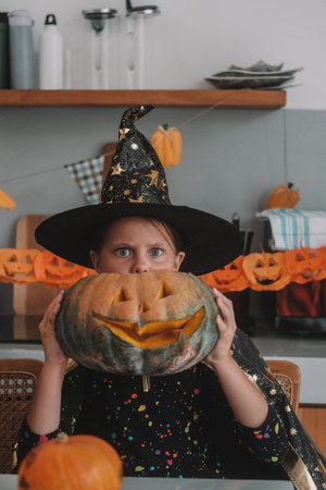 A portrait of a little girl dressed as a witch, holding a large pumpkin with a scary smile carved...