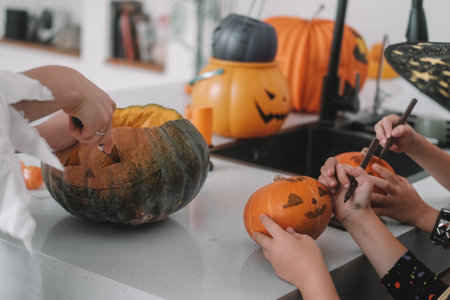 A mother, son and daughter are carving Halloween pumpkins in decorated kitchen. The children are playing and scaring each other. The girl is dressed as a witch and her brother is dressed as a pirate.の写真素材