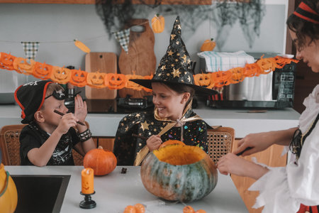 A mother, son and daughter are carving Halloween pumpkins in decorated kitchen. The children are playing and scaring each other. The girl is dressed as a witch and her brother is dressed as a pirate.の写真素材