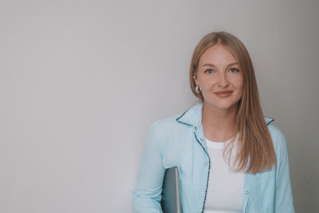A portrait of a blonde businesswoman in a classic shirt standing in an office against a white wall,...