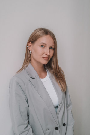 A portrait of a blonde businesswoman in a classic gray jacket standing against a white wall, looking...