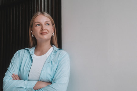 A portrait of a blonde businesswoman in a classic shirt standing in an office against a white wall,...