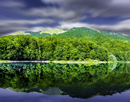 Beautiful landscape of mountain lake on sunny summer day with speedy clouds on blue sky, green forest and calm water mirroring forest, sky and clouds.の写真素材