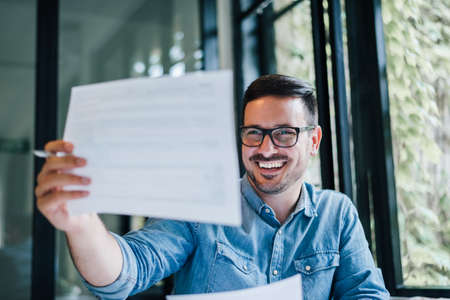 Portrait of young smiling cheerful man entrepreneur or businessman in casual office working with charts and graphs looking at paper documents accounting report spreadsheet happy with the resultsの写真素材