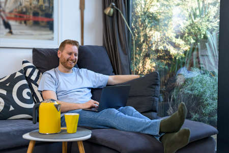 Cheerful smiling young adult man comfortably sitting at home on large sofa using laptop computer spending free leisure time surfing internet web exploring interesting things gathering new knowledgeの写真素材