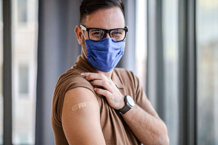 Young adult man showing pointing to bandage on his arm shoulder wearing protective mask done with...