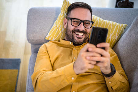 Cheerful young adult mature man happy smiling using smart phone resting lying down on sofa couch at home reading messages on mobile phone Guy with eyeglasses relaxing at home surfing net social mediaの写真素材